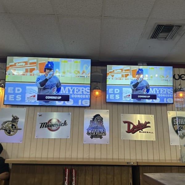 Two large screens show a baseball player in a Dodgers uniform above a bar with various logos.