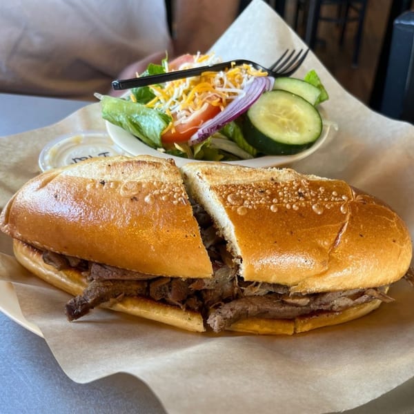 A sliced roast beef sandwich on a bun with a side salad of lettuce, tomato, cucumber, and shredded cheese.