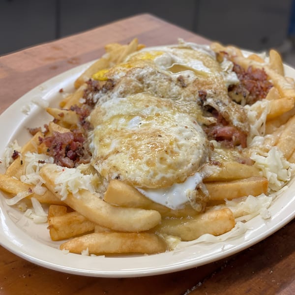 A close-up of a plate of golden french fries topped with fried eggs, bacon bits, and shredded cheese.