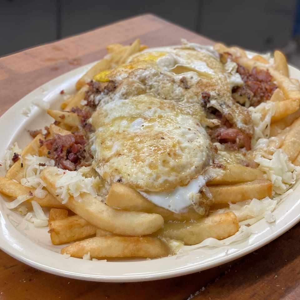 A close-up of a plate of golden french fries topped with fried eggs, bacon bits, and shredded cheese.